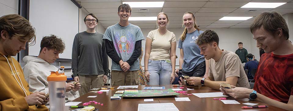 Four students standing behind students playing board game