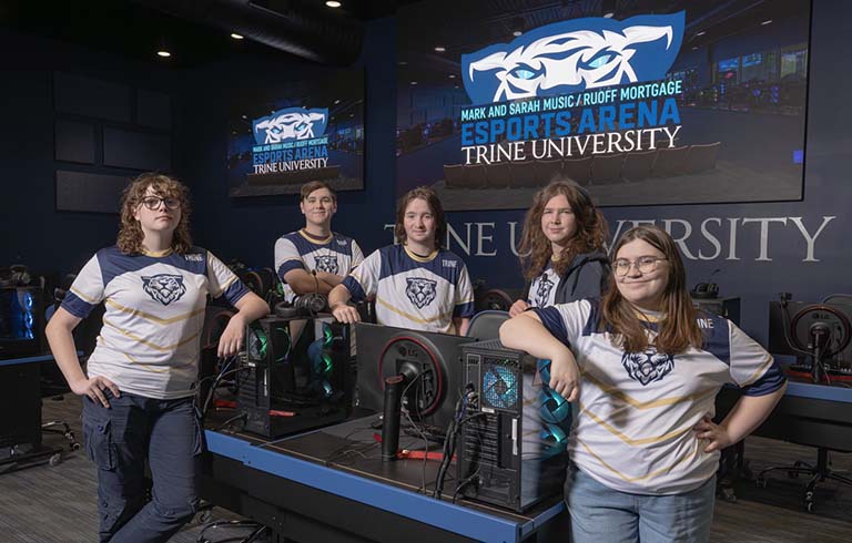 Students leaning on computer towers, standing in front of large gaming screens