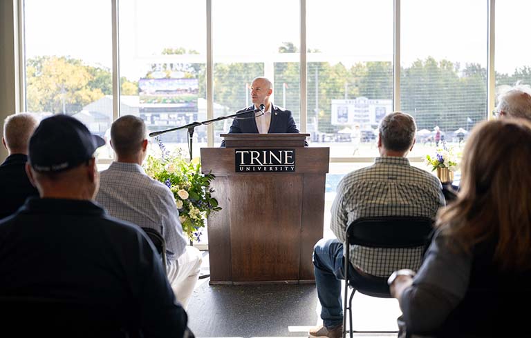 Dr. Kline at podium speaking to crowd; football field is outside windows behind him